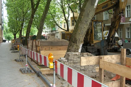 Straßenbaustelle mit Bagger und rot-weißen Absperrungen entlang einer Allee